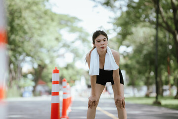 A fit woman cools down after an intense outdoor workout, wiping sweat with a towel. She embodies...