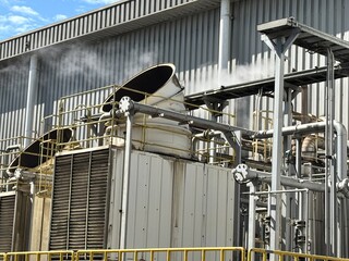 Industrial cooling tower system emitting vapor under blue sky. Heavy-duty piping, valves, and heat exchangers in a manufacturing or refinery plant exterior.