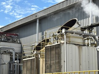 Industrial cooling tower system emitting vapor under blue sky. Heavy-duty piping, valves, and heat exchangers in a manufacturing or refinery plant exterior.