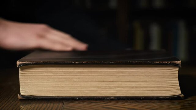 Hand rests on a thick, old book, ring visible, dark background