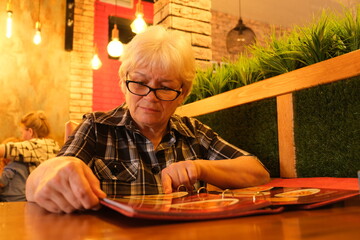 Lunch choice. Caucasian senior woman making an order in the cafe using menu. Soft twilight in a cafe