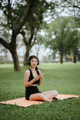 Young Asian woman sits in a quiet park. She wears headphones, enjoys music, practices mindfulness, exercises and digital health with her smartphone.