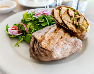 A grilled Chicken Caesar wrap rests on a rustic café table, its golden tortilla folded tight with greens and grilled meat, beside a fresh side salad, background softly blurred in warm light.