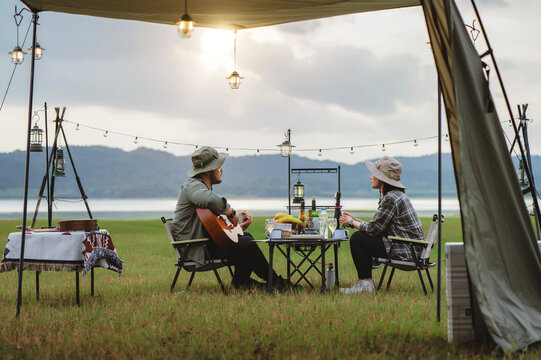 Romantic Asian couple camping with guitar near lake, Man playing guitar for women during lakeside camp, Nature retreat with partner, Romantic melody moment during lakeside retreat - Powered by Adobe