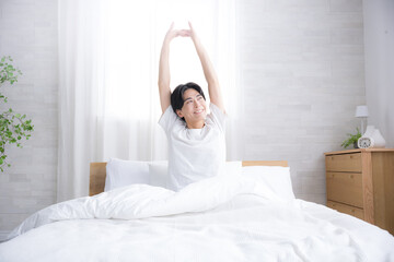 朝日を浴びるベッドで寝起きの若い男性　腕を伸ばしストレッチをする