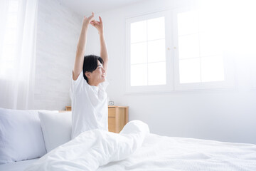 朝日を浴びるベッドで寝起きの若い男性　腕を伸ばしストレッチをする