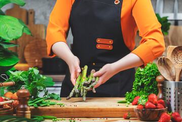 Chef woman in black apron skillfully handles fresh asparagus as she prepares a healthy vegan dish in a lively kitchen.