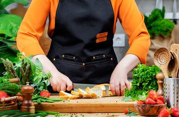 Chef woman in a black apron skillfully slices baguette bread on a cutting board surrounded by fresh greens and healthy ingredients in a cozy kitchen.
