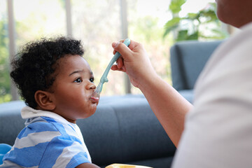 A little boy is eating food that his mother has fed him in the living room at home. Preschool child development. Food menu for 1 year old baby.