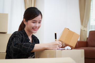 Beautiful Asian female entrepreneur doing online business, writing product information on a parcel box to send to customers. Internet service. Online business idea.