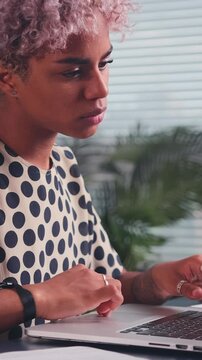 African American woman in a polka-dotted outfit concentrates on her laptop at a workspace. The setting features green plants and a stack of books, creating an engaging environment for productivity.