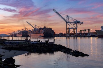 Oakland, USA: People relax on the shore as a tugboat guides container ship into port at sunset....