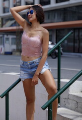 A young Asian woman leans on outdoor stair railing, dressed in a stylish pink top and denim shorts. She wears trendy sunglasses, exuding a relaxed yet fashionable vibe