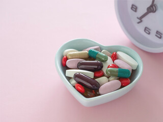 Close up blue heart shaped bowl with various colored dietary supplement pills and clock on pink background. Health supplement concept. Top view with copy space.
