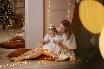 Young Caucasian mother in white dress claps hands, baby girl sits on floor playing. Front view. Cozy kids room with wooden floor,pillows, curtain tent. Concepts: bonding, childhood, play, care.