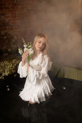 Young Caucasian woman, mid-20s, light brown hair, in white dress, front view, sits in shallow artificial pond with fog and flowers, brick wall behind. Concepts: fantasy, nature, calm, dreamlike.