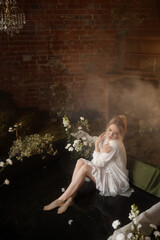 Young Caucasian woman, mid-20s, light brown hair, in white dress, front view, sits in shallow artificial pond with fog and flowers, brick wall behind. Concepts: fantasy, nature, calm, dreamlike.
