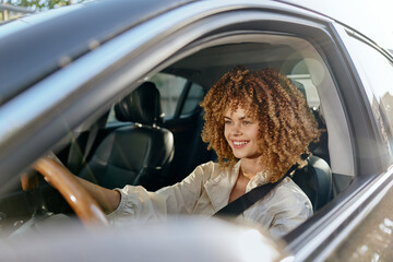 Smiling woman with curly hair driving a modern car, wearing a beige blouse, enjoying a sunny day and safety with seatbelt fastened, casual lifestyle.