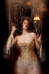 Elegant young circus actress in golden corset and gloves, standing under chandelier, front view. Retro theatrical look for circus, burlesque, performance, vintage stage shows.