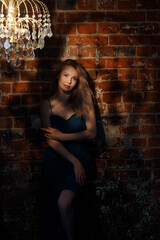 Young Caucasian woman in blue dress stands in dramatic light, arms crossed. Front view. Brick wall with chandelier in vintage room. Concepts: mood, elegance, solitude, mystery.