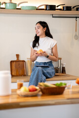 Smiling asian woman holding cookies bowl looking away from tablet while sitting on kitchen counter.