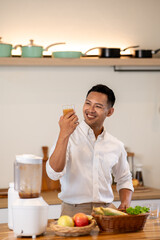 Smart asian man looking at a glass of protein smoothie after making drink at kitchen wooden counter.