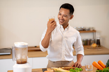 Happy asian man looking at a glass of protein smoothie after making drink at kitchen wooden counter.