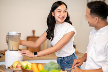 Asian woman using blender and smiling at man holding knife making smoothie drink at kitchen counter.