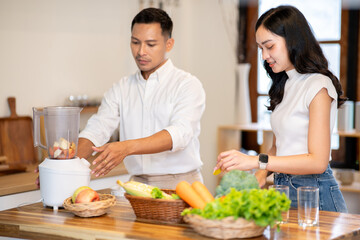 Asian man using blender while woman holding apple cuts making smoothie drink at kitchen wood counter