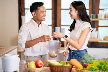 Asian man holding blender and woman putting in apple cuts making smoothie drink at kitchen counter.