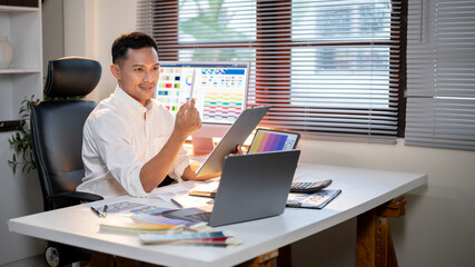 smiling asian man designer holding clipboard looking at stylus while sits at office's working table.