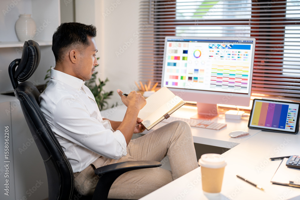 Aufkleber asian designer man looking at book while sitting with computer and tablet on working table in office