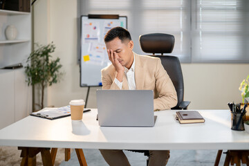 Asian businessman with a hand covering face looking tired working too long on laptop at office table