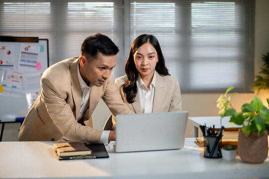 Asian businessman looking closely at female coworker's laptop screen on working table in the office.