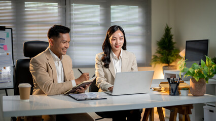 Asian businessman writing in notebook female coworker looking at laptop while sit at table in office