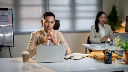 Asian businessman joining hands and thinking while sitting at working table aside female coworker in office.