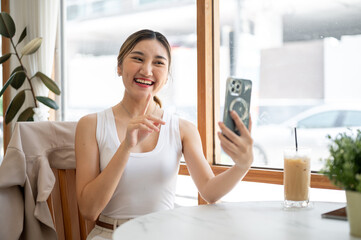 Asian woman holding phone talking on video call with finger pointing at cafe table with iced coffee.