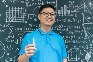 Smiling Asian male teacher holding chalk in front of blackboard with math formulas, education and classroom concept