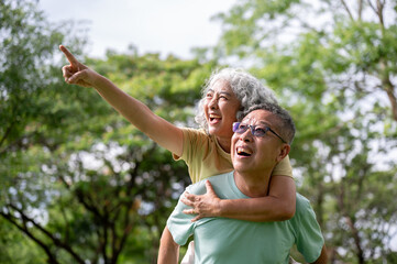 Glasses old man carrying old woman on his back and she's pointing away while standing in the park.