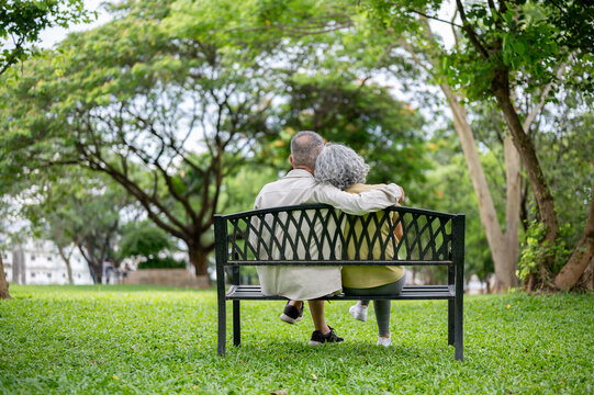 Back view of old man hugging a woman shoulder as sitting leaning against each other on bench in park