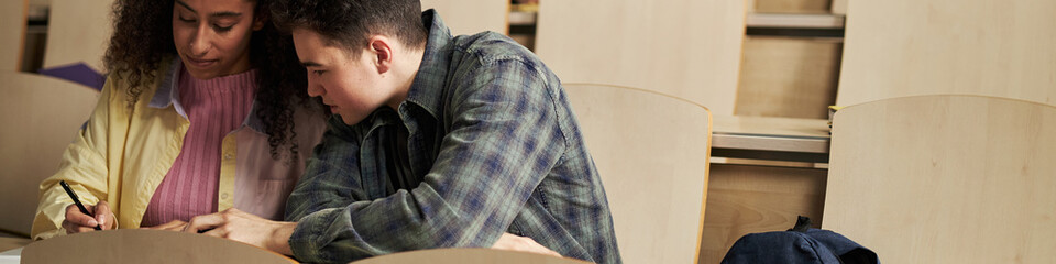 Fototapeta premium Header of young Caucasian man and Black woman sitting together in university lecture hall collaborating on academic assignment, both focused on writing in notebook