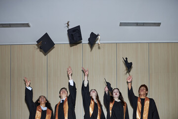 Diverse group of university young students wearing graduation gowns and tossing caps into air,...