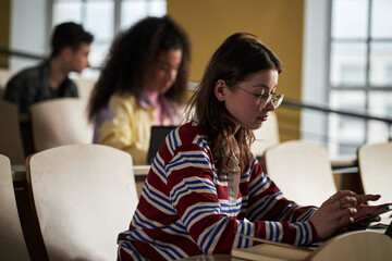 Caucasian young woman sitting in lecture hall using digital tablet, multiethnic university students...