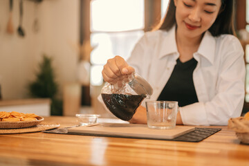 Close-up of a woman pouring a black coffee from a glass pot into a clear cup on a wooden kitchen...