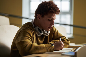Teenage Black man sitting at desk writing in notebook with over ear headphones around neck in university classroom setting, focused on studying and taking notes during lecture