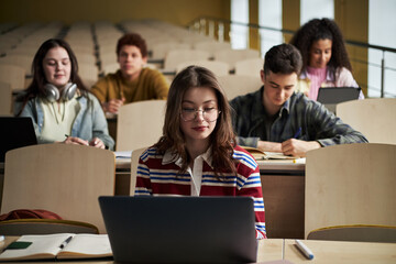 Group of multiethnic university students studying in lecture hall, young Caucasian woman with glasses using laptop in foreground, teenagers and young adults writing notes and working on laptops