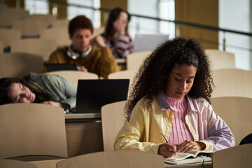 Young adult Black woman writing in notebook while sitting in lecture hall, multiethnic group of university students in background using laptops and resting during class