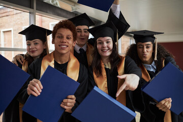 Group of multiethnic teenagers wearing graduation caps and gowns celebrating university...