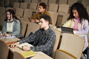 Group of multiethnic university students sitting in lecture hall taking notes and using digital tablet, listening to professor explaining new material, taking notes