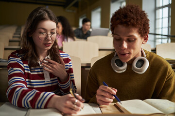 Caucasian young woman and Black man studying together in university lecture hall, writing notes in...